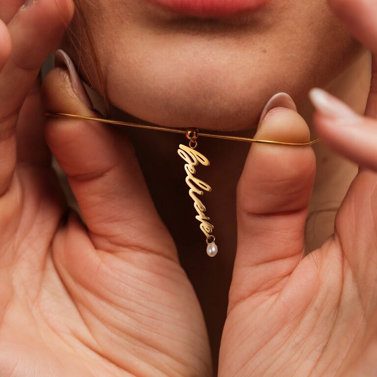 "Believe-Charm" goldener Anhänger mit Perlen, mit einem kursiven „Believe“-Design auf einem sanften Hintergrund.