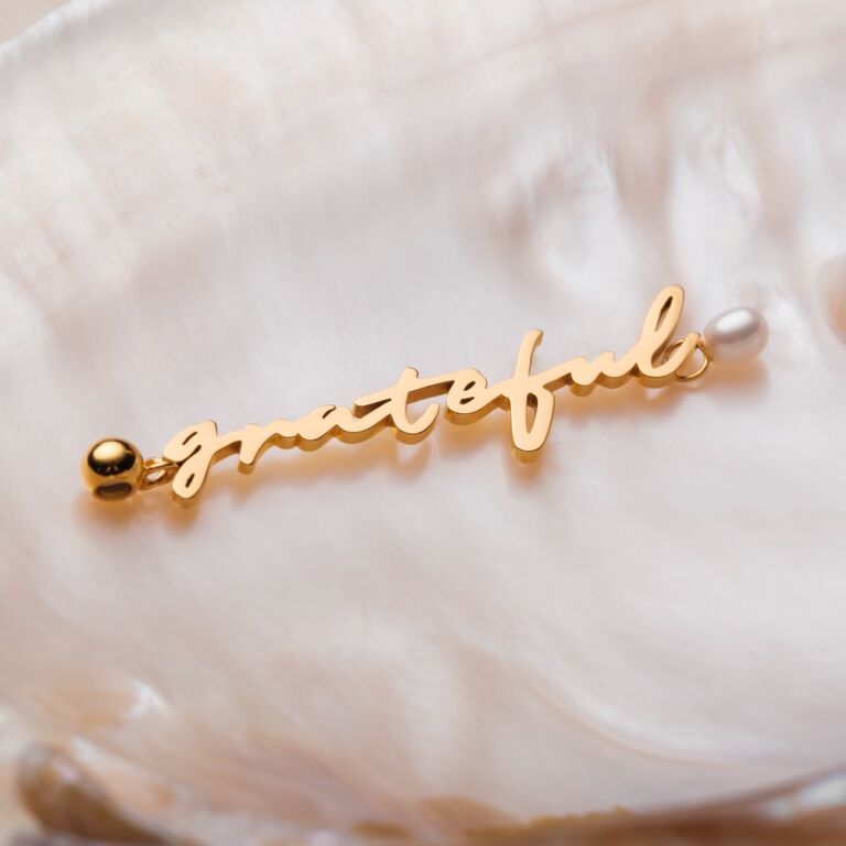 "Grateful-Charm"-Anhänger in Gold mit Perlen, mit eleganter Schrift auf strukturiertem Muschelhintergrund.
